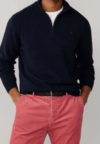 Homme portant un pull bleu marine à fermeture éclair quart, par-dessus une chemise blanche, pantalon rose avec ceinture noire, mains dans les poches, sur fond gris uni.