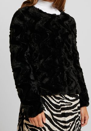 Personne portant une veste en fausse fourrure noire texturée par-dessus un col roulé blanc et une jupe imprimée zèbre noire et blanche.