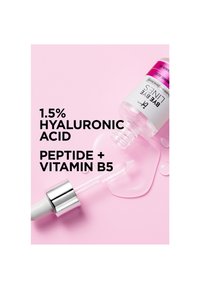 Flacon de sérum étiqueté "1,5 % d'acide hyaluronique, peptide + vitamine B5" avec compte-gouttes et liquide renversé sur fond rose.
