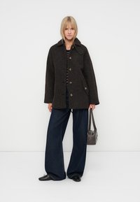 Palton din lână maro cu nasturi mari, guler cu fante, și buzunare laterale, asociat cu pantaloni din denim închis, cu croi larg, și o geantă mică gri.