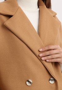 Mujer ajustando la solapa de un abrigo de lana de color camello con doble botonadura sobre un suéter de cuello alto blanco de canalé.