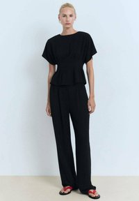 La modella è in piedi contro uno sfondo azzurro chiaro, indossando un top peplum nero e pantaloni neri, abbinati a sandali rossi.