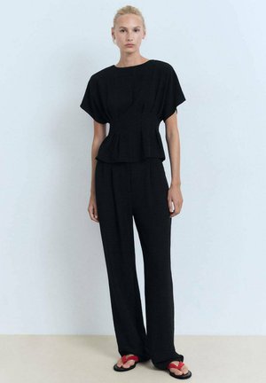 La modella è in piedi contro uno sfondo azzurro chiaro, indossando un top peplum nero e pantaloni neri, abbinati a sandali rossi.
