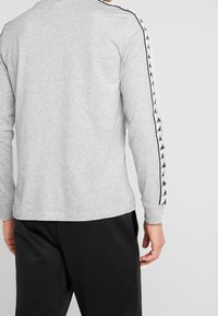 Šedá dlouhá trička s černými logovými akcenty podél rukávů, s kulatým výstřihem a hladkým bavlněným materiálem.