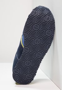 Sole z námořnické modré barvy s hexagonálním vzorem, vyrobená z texturované černé gumy, s žlutým akcentem blízko horní hrany.