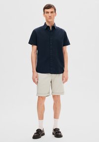 Kortärmad marinblå skjorta med knappar, tillsammans med ljusbeige rullade shorts, bärs med svarta skor och vita strumpor, ståendes mot en neutral bakgrund.