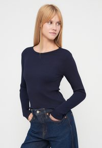 Bluză navy cu mâneci lungi și dungi, cu decolteu la baza gâtului, asortată cu blugi din denim cu talie înaltă. Țesătura are o textură fină.