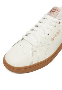 Biela teniska Reebok s béžovým logom, bielymi šnúrkami a podrážkou v prírodnej farbe, s vyšitými detailmi a perforáciami na oblasti špičky.