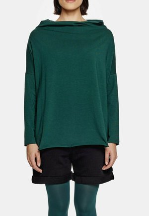 Kobieta ubrana w luźną, ciemnozieloną bluzkę z długim rękawem, czarne podwinięte szorty i turkusowe rajstopy, stojąca na białym tle.