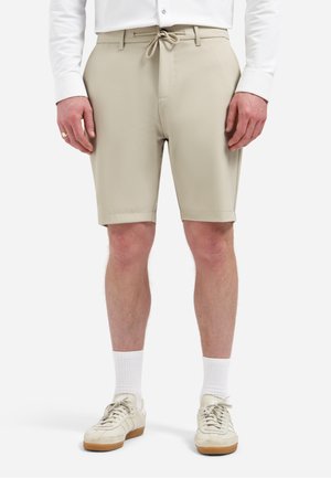Man draagt beige korte broek tot op de knieën met trekkoord, wit shirt met lange mouwen, witte sokken en crème-kleurige sneakers.