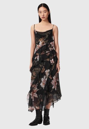 Kobieta w czarnej sukience midi w kwiaty bez rękawów z warstwowymi falbanami i czarnymi botkami, stojąca na tle jednolitego tła.