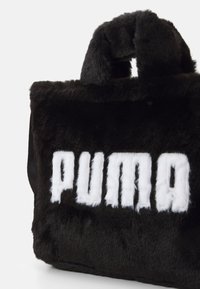 Sort faux fur taske med en blød tekstur, der har et fremtrædende hvidt "PUMA" logo. Den har et kort, fyldigt håndtag. Kompakt rektangulær form.