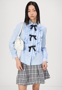 Light blue shirt featuring ruffled front detail and black bow accents, paired with a gray plaid skirt and a small white handbag.