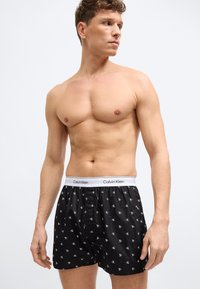 Άνδρας χωρίς πουκάμισο φοράει μαύρα μποξεράκια Calvin Klein με λευκή ελαστική ζώνη μέσης που φέρει το λογότυπο και μικρά λευκά μοτίβα "cK", κοιτάζοντας προς τα πλάγια.