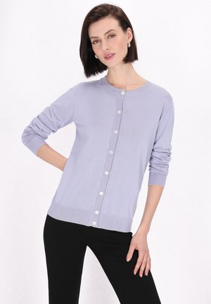 Cardigan lila deschis cu guler rotund, mâneci lungi și țesătură texturată. Prezentă o închidere cu nasturi și manșete cu nervuri, purtat peste pantaloni negri.