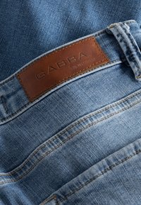 Světle modré džínové kalhoty s hnědým koženým logem "GABBA" na pásku. Viditelné detaily švů a textura.