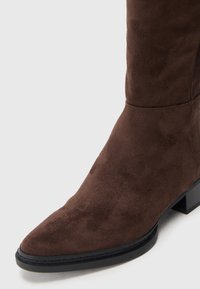 Botín de ante marrón hasta el tobillo con puntera afilada, tacón cuadrado y textura suave. La costura en la parte superior añade un sutil interés de diseño.