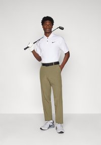 Bijela Nike polo majica s tri gumba i crnim logotipom, maslinaste hlače, crni pojas i sivi Nike golf cipele. Drži golf štap preko ramena.