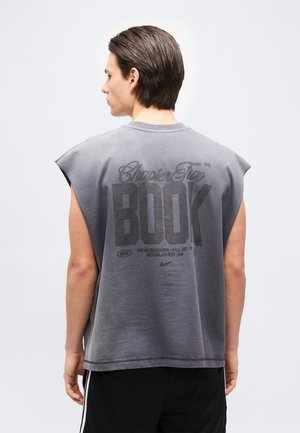 Junger Mann seitlich stehend, trägt ein ärmelloses graues Shirt mit dem Text „Chapter Two BOOK“ auf dem Rücken, kombiniert mit schwarzen Shorts mit weißen Streifen.