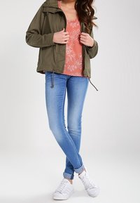 Veste légère vert olive, haut à fleurs corail, jean skinny bleu clair et baskets blanches. La veste est équipée d'une capuche et d'un cordon de serrage.