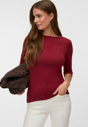 Vrouw met lang bruin haar die een bordeauxrode driekwart mouw top en witte broek draagt, en een opgevouwen donkerbruine jas vasthoudt.