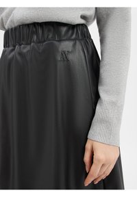 Jupe en cuir noir avec une taille élastique, arborant un logo "AX" en relief. La texture est lisse et brillante. Une manche de pull gris à côtes est visible.