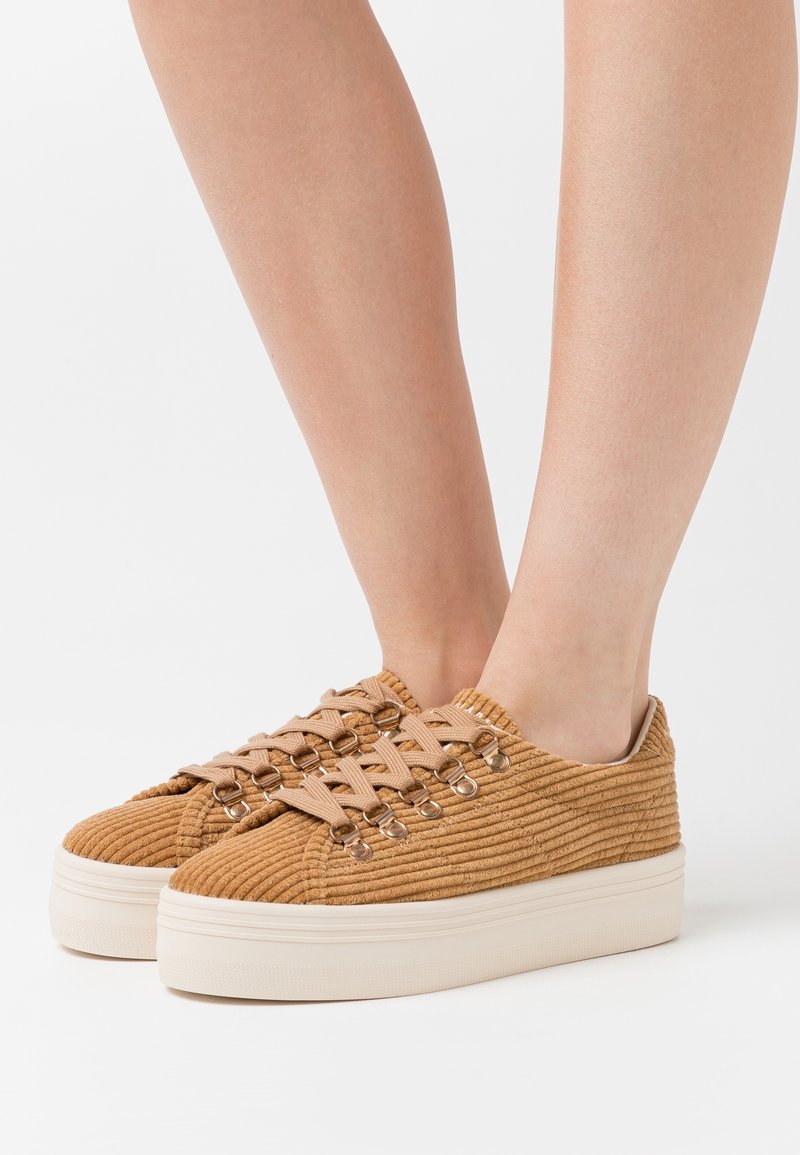 Zapatillas de plataforma de pana en tono marrón con cordones beige, con ojales de metal redondos y parte superior texturizada. Suela de goma plana y diseño de punta redonda.