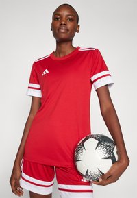 Rode sportieve trui met witte strepen op de mouwen, gecombineerd met bijpassende shorts, en een zwart-wit gestreepte voetbals bal vasthoudend.