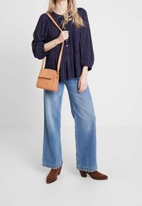 Blusa blu navy con pois bianchi, con bottoni anteriori, abbinata a jeans a gamba larga blu e stivaletti alla caviglia in suede marrone. Borsa a tracolla marrone.