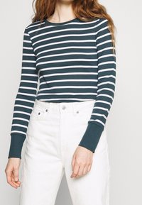 Långärmad skjorta i mörk teal med vita horisontella ränder. Har rund halsringning och åtsittande ärmslut, i kombination med vita högmidjade byxor.