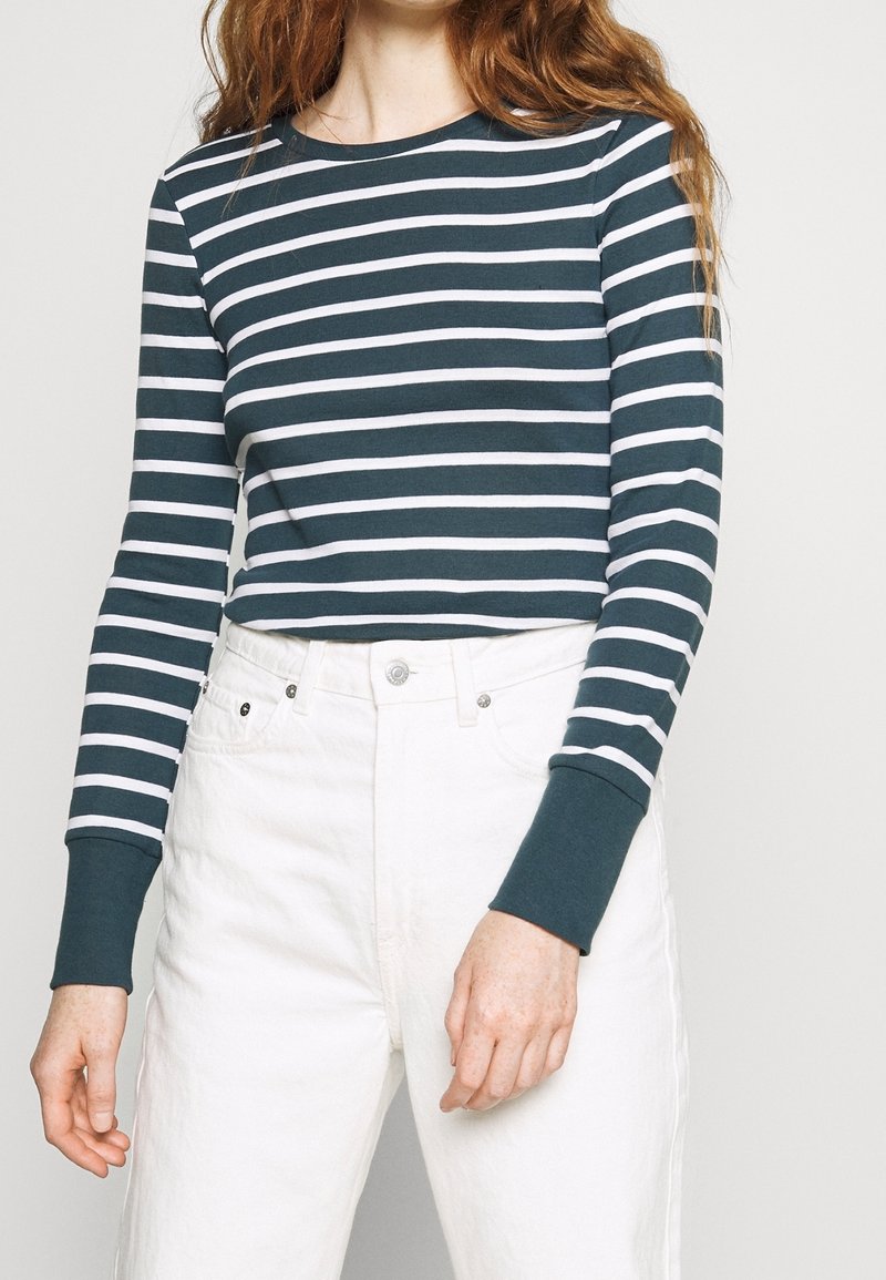 Långärmad skjorta i mörk teal med vita horisontella ränder. Har rund halsringning och åtsittande ärmslut, i kombination med vita högmidjade byxor.