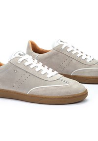 Zapatillas de ante en gris claro con cordones blancos. Punta redondeada, suela de goma marrón y detalles perforados en los laterales, con sutiles acentos de logo.
