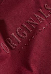 Sweat-shirt bordeaux avec des lettres texturées épelant "ORIGINALS" et le nom de la marque "JACK & JONES" en dessous, présentant un col rond.