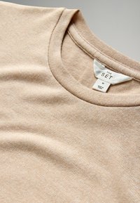 Beige T-shirt met korte mouwen en een ronde hals; de stof heeft een zachte, gemêleerde textuur. Het label aan de binnenkant zegt "THE SET" en maat "M."