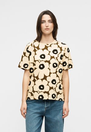 Giovane donna che indossa una maglietta a maniche corte beige e marrone con stampa floreale e jeans blu, in piedi contro uno sfondo chiaro e uniforme.