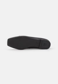 Chaussure plate noire avec une tige en cuir lisse, un bout carré et une semelle en caoutchouc texturée avec un marquage sur le talon.