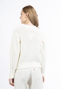 Pull en tricot blanc avec des motifs texturés, des poignets et un ourlet côtelés, et des détails complexes sur les manches. La vue arrière montre un tissage ouvert.