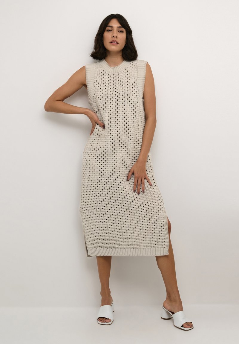 Beige gestricktes ärmelloses Kleid mit einem strukturierten geometrischen Muster, runder Ausschnitt und Seitenschlitzen. Kombiniert mit weißen offenen Mules.