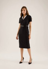 Vestido preto justo com mangas curtas, decote em V, colarinho branco em contraste e um cinto justo na cintura. Tecido suave e design até ao joelho.