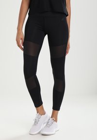 Leggings atletici neri con dettagli a pannello trasparente, design aderente e texture liscia, abbinati a scarpe da ginnastica di colore chiaro.
