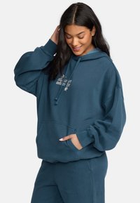 Oversized teal hoodie made of soft fabric, featuring a front pocket, drawstring hood, and embroidered text detail.