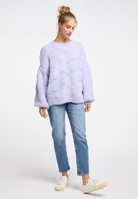 Lila gestrickter Pullover mit großen, strukturierten Wellenmustern, Ballonärmeln und rundem Ausschnitt, kombiniert mit blauen Slim-Fit-Jeans und weißen Sneakern.