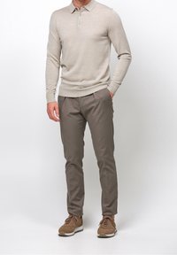 Polo beige à manches longues avec patte de boutonnage à trois boutons, associé à un pantalon marron slim et des baskets marron à semelles blanches, se tenant sur un fond uni.