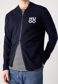 Marinblå zip-up jacka med ribbad krage och mudd, med en vit "HUGO" logotyp på vänster bröst, lagd  över enrandig skjorta.