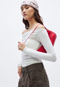 Witte off-shoulder top met plooidetail, gecombineerd met donkerbruine hoge taille broek. Houdt een kleine rode tas met een dunne band vast.