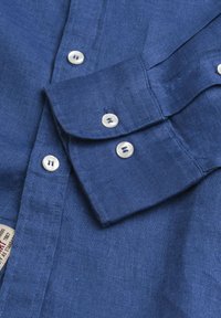 Camicia in lino blu con colletto a punta, polsini abbottonati e bottoni bianchi. Il tessuto presenta una finitura strutturata e leggermente stropicciata.