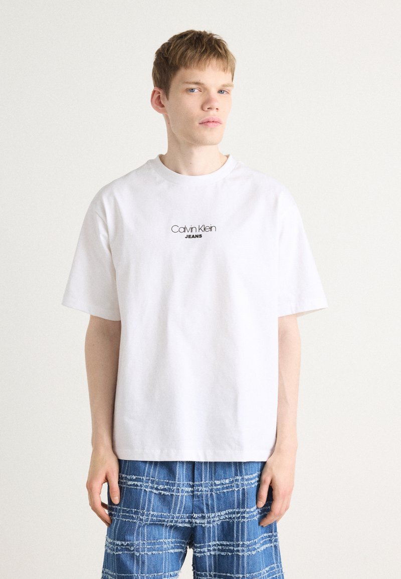 Camiseta blanca de algodón con cuello redondo y mangas cortas, con un logo negro de "Calvin Klein JEANS" en el pecho. Combinada con pantalones azules estampados.
