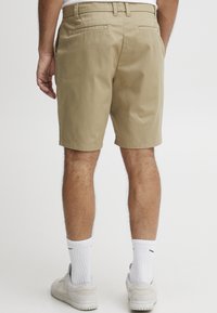 Beige Shorts aus glattem Baumwollmaterial mit zwei Gesäßtaschen und einem taillierten Bund. Getragen mit weißen Socken und hellen Sneakers.