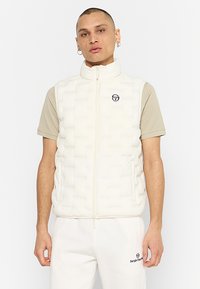 Chaleco acolchado blanco con cremallera frontal, bolsillos laterales y un detalle de logo en el pecho. Llevado sobre una camisa de manga corta beige.