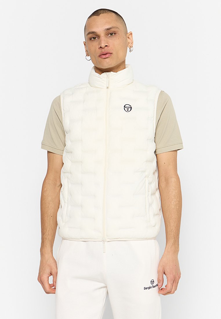 Chaleco acolchado blanco con cremallera frontal, bolsillos laterales y un detalle de logo en el pecho. Llevado sobre una camisa de manga corta beige.